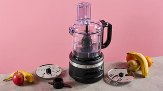 A matte black KitchenAid 7 cup food processor is pictured against a pink background. It is sitting on a grey stone-effect surface on which also sites the reversible slicing and grating disc on the left, with the plastic dough hook positioned just in front, and to the right, the thick slice disc that is only available with the KFP0719 configuration.